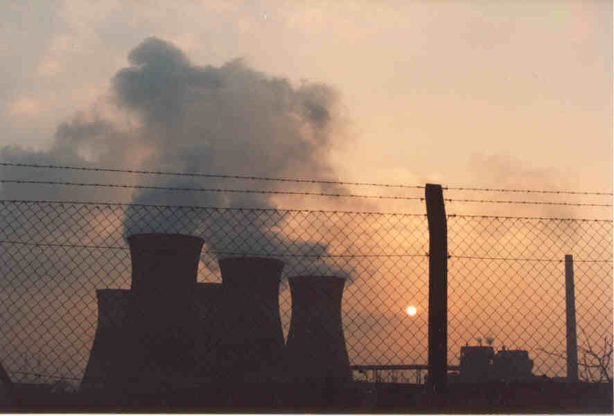 Taken through chain link fencing, the sun is in the background, almost lost in the haze of the power station